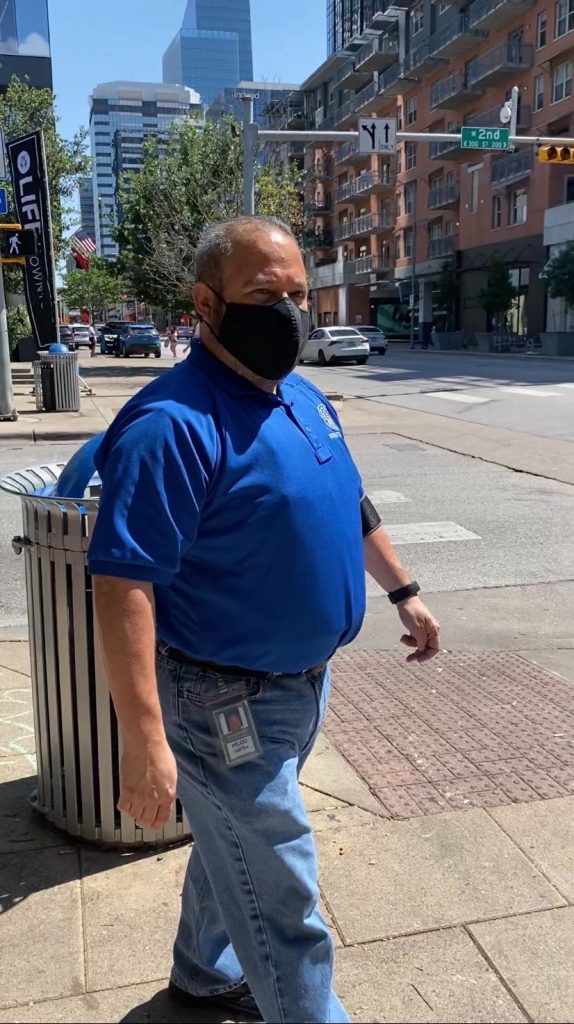 A picture of a balding white man in a royal blue polo with a black face mask standing on the corner of a sidewalk