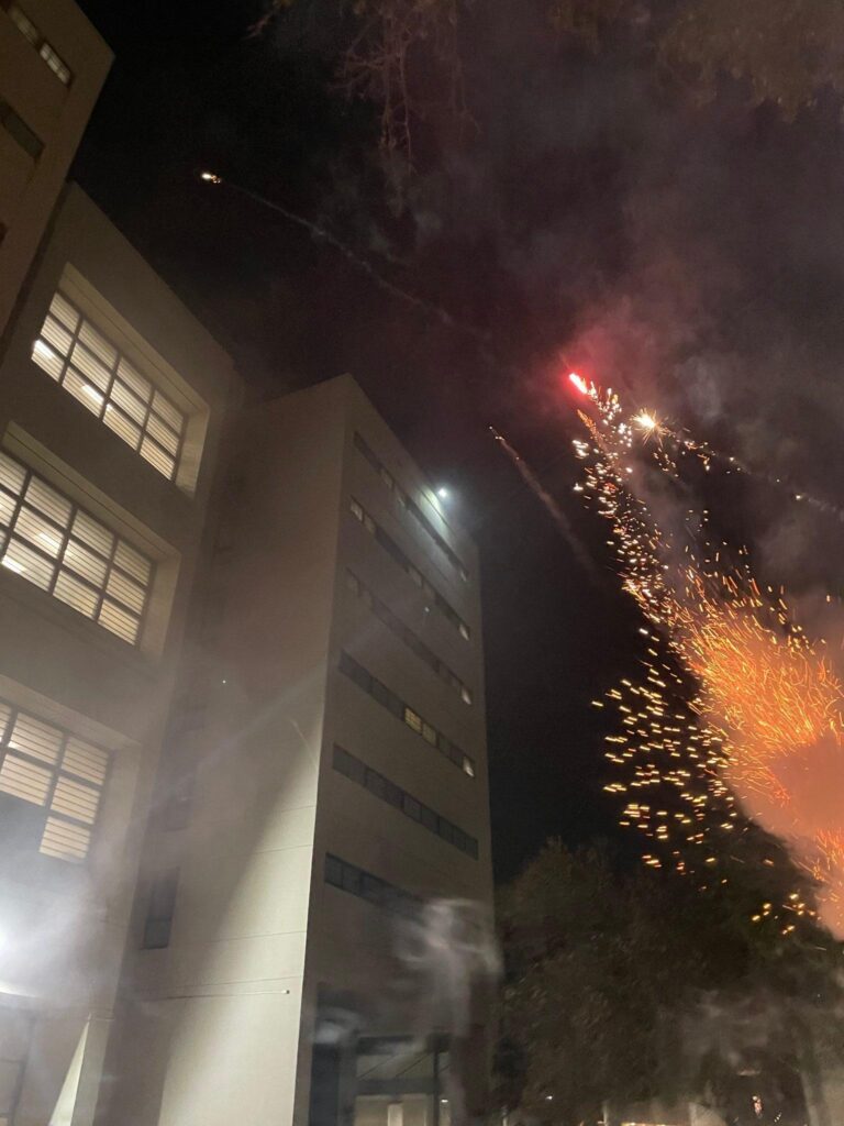 A grey concrete building on the left. An orange burst from a firework approaching the building from the right.