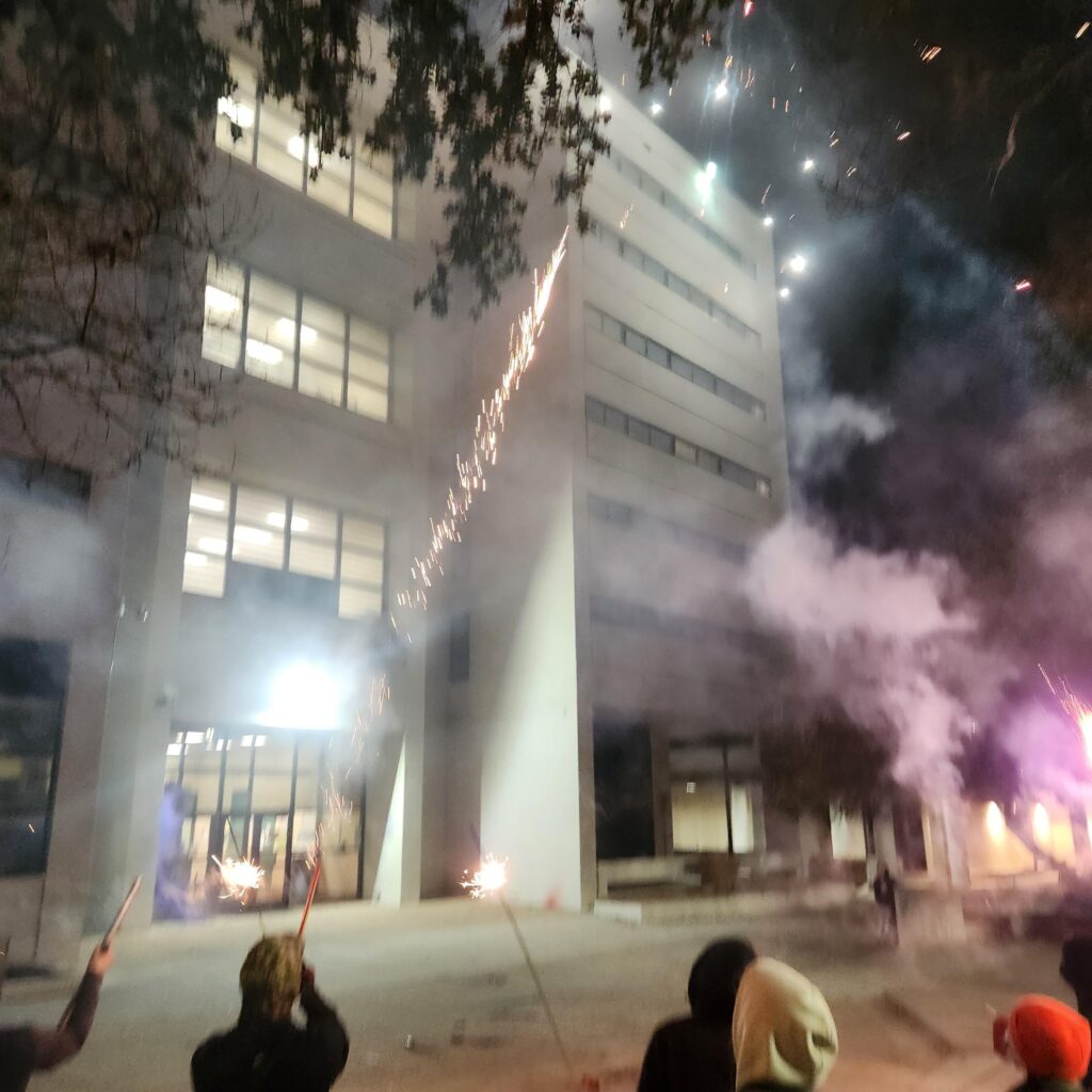 A crowd in front of the concrete building of the jail with fireworks and smoke in the air