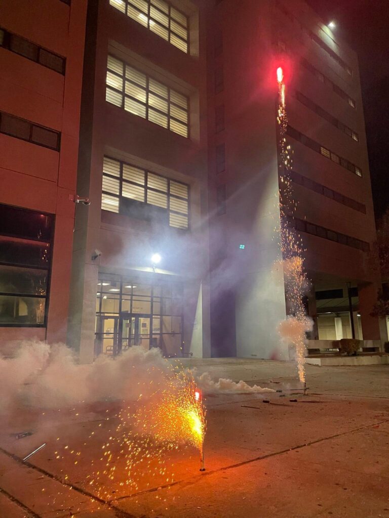Two fireworks launching from in front of the jail. One is orange, the other is yellow, both launching from canisters on the ground.