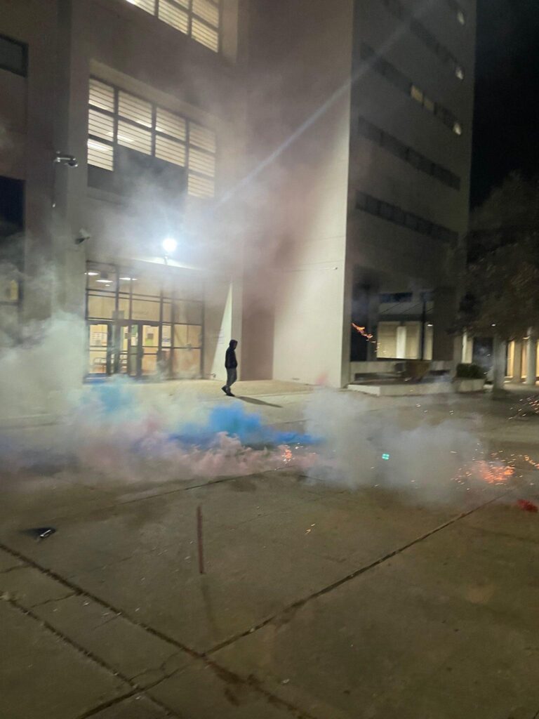 Smoke from smoke bombs obscuring the front of the jail, a large concrete building in the background of the photo. The smoke is white, blue, and pink colored.