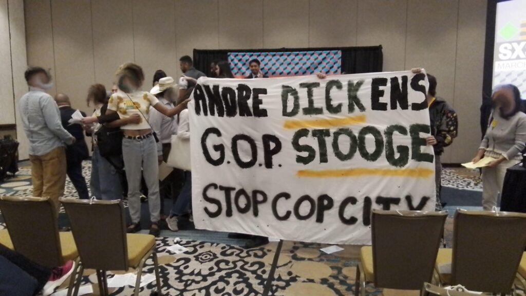 a group of people hold a banner in front of a stage. the banner reads "Andre Dickens, G.O.P. Stooge, Stop Cop City"