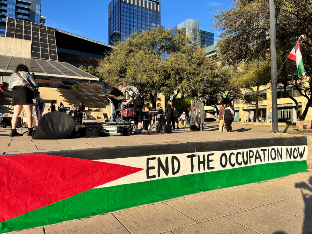 Musicians perform in front of city hall. A mural of a palestinian flag sits on the wall with "End the Occupation Now" written over it.