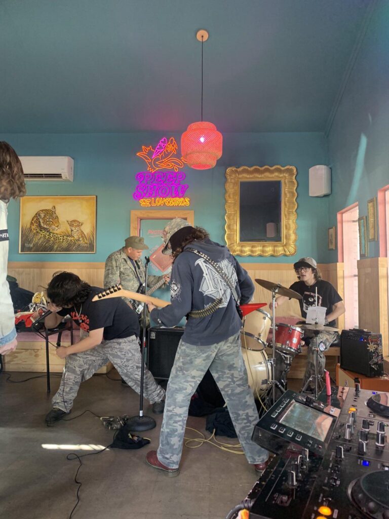 4 members of a band play in a bar. A guitar, basee, and drumset are visible in the photo. The back wall is teal, with two paintings on the back wall and a neon sign that says "Peep Show at Lovebirds"
