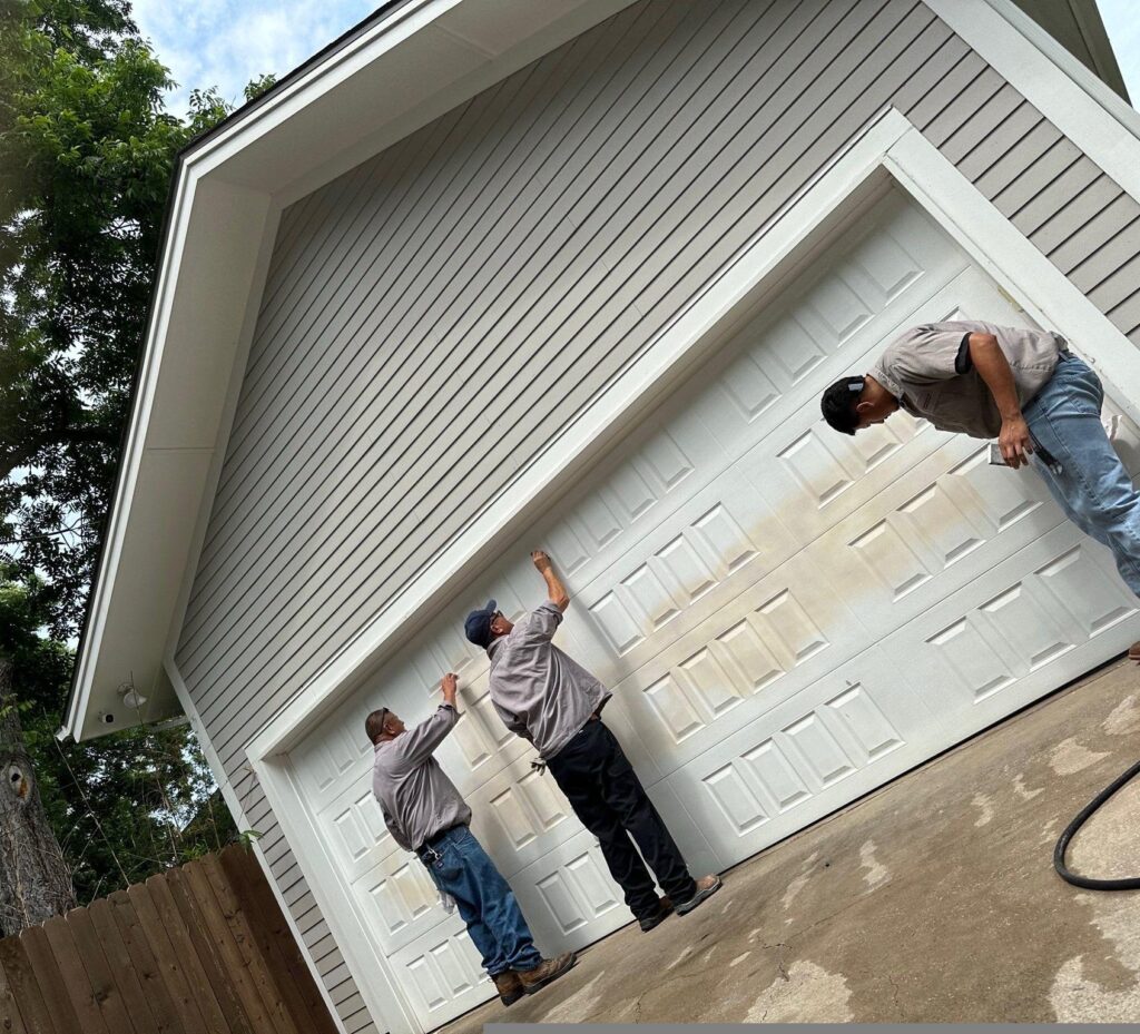 A grey garage with a white garage door. Three painters in grey shirts and jeans stand in front of it. A message written in red paint is barely visible through the new paint on top. 